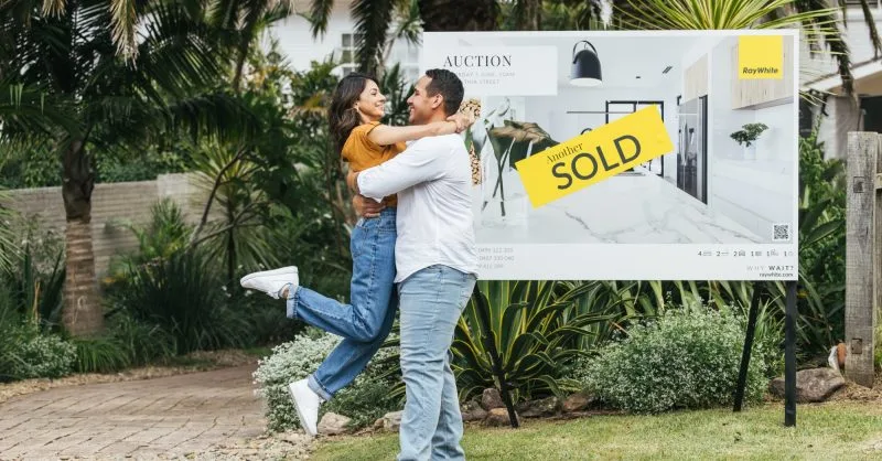 Couple celebrating in front of a sold sign at their first home